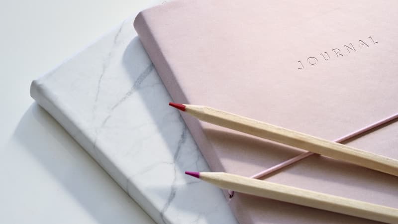 Two journals stacked on top of each other. The bottom one is grey marble pattern and the top one is light pink. There are purple and pink pencils laying on top