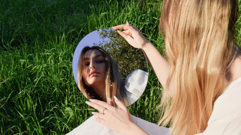 A woman looks into a circular mirror outside on the grass