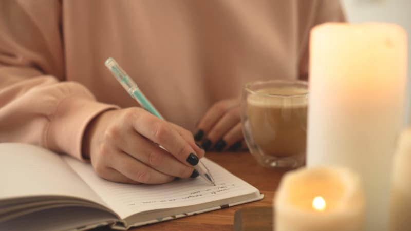 A woman with black nails reflects on self compassion journal prompts in her journal. There is a candle and coffee near her.